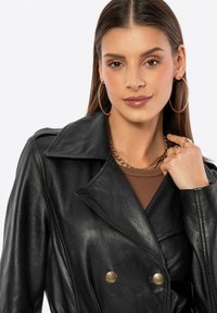 Chaqueta de cuero negra de doble botonadura con cuello puntiagudo y detalles de botones dorados. Llevada sobre una camiseta marrón, combinada con joyería dorada gruesa.