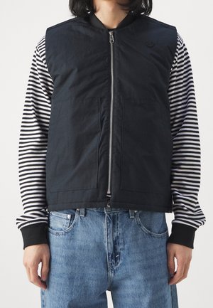 Personne portant un gilet noir zippé sur une chemise à manches longues rayée noir et blanc avec un jean en denim bleu, debout devant un fond uni.