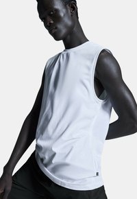 Weißes ärmelloses Sportshirt aus glattem Stoff, mit abgerundetem Saum und seitlichen Nähten. Schwarzes Logotag am unteren Rand sichtbar.