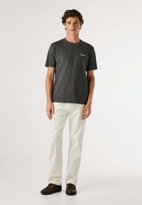 Grijze T-shirt met korte mouwen en ronde hals, voorzien van een klein wit logo op de linkerborst, gecombineerd met lichte broek en bruine schoenen.