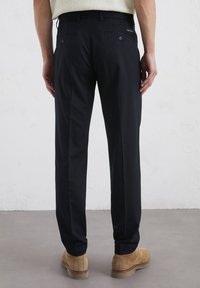 Pantalon noir pour homme avec une texture lisse, coupe ajustée, deux poches arrière avec des boutons et un branding minimal. Porté avec des chaussures marron clair.
