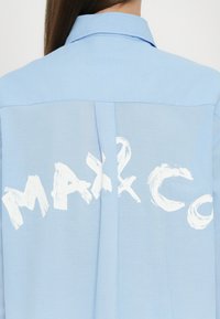 Vaaleansininen paita, jossa on levitetty kaulus ja suuren valkoisen graafisen tekstin "MAX & CO" printti selässä. Silkkinen kangas ja rento malli.