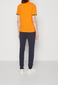 Personne se tenant de dos, portant un polo orange à manches courtes, un pantalon de jogging bleu marine, des baskets blanches et des chaussettes blanches sur un fond uni.