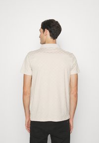 Polo de color beige claro con un patrón geométrico texturizado, de manga corta y con cuello. La tela parece suave y casual. Se llevan pantalones negros.