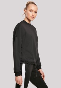 Sudadera negra con cuello redondo, cintura elástica y detalles de cordón. Textura suave y corte relajado; combinada con pantalones negros que tienen franjas blancas.