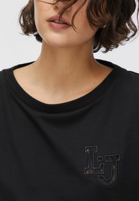 Haut noir à manches courtes avec un col rond et un logo "L.J." brodé en texture contrastée. Tissu doux et lisse.