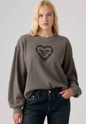 Femme blonde portant un sweat-shirt gris tricoté avec un motif de cœur noir et un jean bleu foncé, debout avec une main dans la poche.