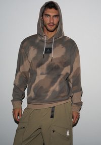 Brązowy hoodie w tie-dye z przednią kieszenią i kapturem ze sznurkiem, w połączeniu z zielonymi spodniami cargo z wieloma kieszeniami i logo.