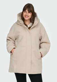 Bež villasegu mantel suure kapuutsiga, üleliia suuruse, küljetaskute ja pehme tekstuuriga, esiseemiga ja minimalistikus disainis.