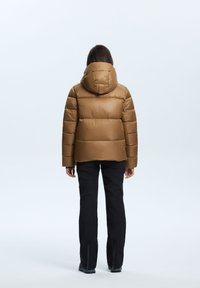 Doudoune brown avec capuche, présentant une texture brillante et un design matelassé. Portée par-dessus un pantalon noir avec des accents de fermeture éclair au niveau des chevilles.