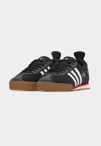 Μαύρα παπούτσια Adidas Samba με λευκές ρίγες, σόλες από καουτσούκ χρώματος τσίχλας, κόκκινα και μπεζ στοιχεία στη μέση της σόλας και χρυσό λογότυπο στις πλευρές και στη γλώσσα.