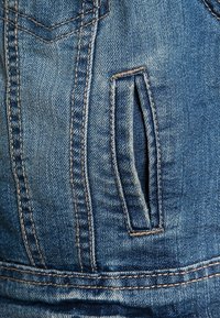 Denimtyg med en bleknad blå nyans som har en liten sidficka. Synlig sömnad i en kontrasterande brun färg ger detalj.