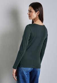 Tummanvihreä pitkähihainen paita pyöreällä kaula-aukolla, valmistettu pehmeästä kankaasta. Yhdistetty sinisiin farkkuihin. Näkyvät ompelutiedot.