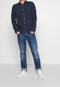 Marineblå skjorte med knaplukning og brystlomme, parret med let slidte blå denimjeans og hvide sneakers. Afslappet pasform, lange ærmer.