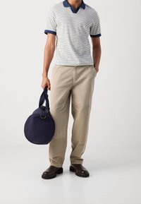 Pruhovaná polokošile v bílé a námořnické barvě, béžové kalhoty a tmavě hnědé boty. Drží kulatou námořnickou tašku s pevným uchem.