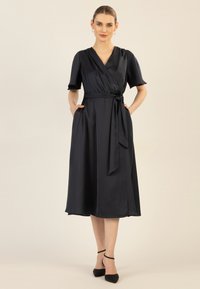Navy wrap dress in satiinimateriaalista, jossa on lyhyet hihat, sivutaskut ja vyö, virtaava siluetti ja polven alapuolelle ulottuva pituus.