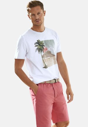 Weißes Baumwoll-T-Shirt mit Palmenmotiv, kombiniert mit rosa Shorts und einem geflochtenen Gürtel, vor einem einfarbigen Hintergrund stehend.