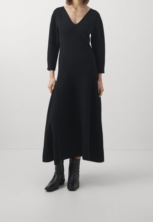 Femme portant une longue robe noire à col en V avec des manches trois-quarts et des bottines noires, debout devant un fond clair uni.