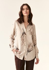 Camisa abotonada de un beige claro con patrones de hojas en marrón oscuro, de manga larga y corte relajado. La tela parece suave y ligeramente texturizada.
