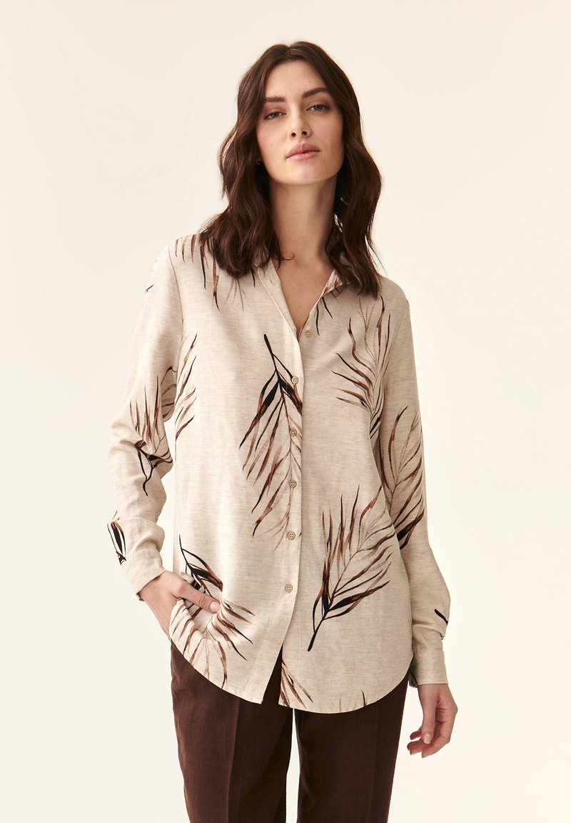 Camisa abotonada de un beige claro con patrones de hojas en marrón oscuro, de manga larga y corte relajado. La tela parece suave y ligeramente texturizada.