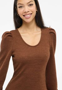 Ruskea, pitkähihainen toppi, jossa on uritettu kangas, pyöreä pääntie ja lievästi pussimaiset olkapääkoristeet, valmistettu teksturoidusta, pehmeästä kankaasta.