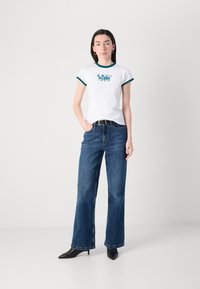 White cotton t-shirt with green ribbed neckline and logo print, paired with dark blue wide-leg jeans and black pointed-toe heels.