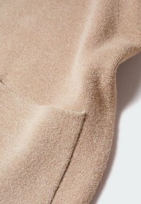 Tissu en laine beige doux avec une couture visible et une texture subtile, montrant des plis de tissu et un tombé naturel.