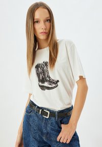 Hvid T-shirt med et grafisk motiv af sorte cowboy støvler med pink detaljer. Kombination med blå denim jeans og et sort bælte med nitter.