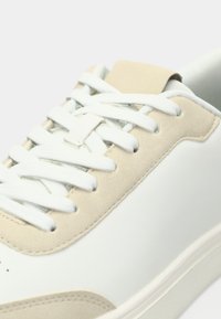 Αθλητικό παπούτσι με λείο λευκό δερμάτινο επάνω μέρος, μπεζ σουέντ λεπτομέρειες, επίπεδα λευκά κορδόνια και επενδεδυμένο κολάρο. Μινιμαλιστικός σχεδιασμός, λαστιχένια σόλα.