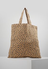 Beige totebag med svart oregelbundet prickmönster, stående upprätt på en vit yta mot en enkel grå bakgrund.