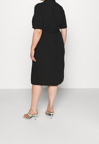 Nainen, jolla on päällään musta polvipituinen mekko, jossa on lyhyet hihat ja vyö, näkyy selkeästä takaa, seisten yksinkertaista vaaleaa taustaa vasten.