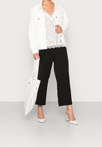 Žena v bílém saku, krajkovém topu, černých širokých kalhotách a krémových lodičkách, držící bílou a béžovou pruhovanou tašku, stojící na šedé podlaze.