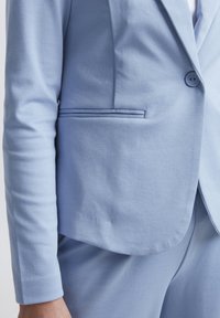 Světle modrý saka na míru s texturovanou tkaninou, zapínání na jeden knoflík a slim fit. Obsahuje náprsní kapsu a jednoduchý design klopy.