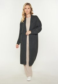 Cardigan lung, cu deschidere frontală, realizat dintr-un material texturat de culoare gri închis, având mâneci lungi și o croială lejeră. Purtați deasupra unei ținute bej.