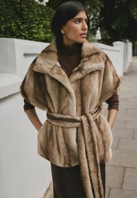 Faux fur vest in light brown with a belted waist, large collar, and textured finish. Worn over a dark turtleneck.