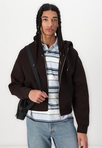 Sweat à capuche marron avec une grande capuche, porté sur un polo rayé bleu clair. Modèle tenant un petit sac noir, se tenant devant un fond blanc.