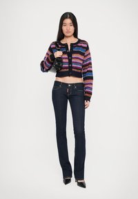 Cárdigan de punto, corto, con rayas horizontales multicolores, combinado con jeans acampanados de color azul oscuro y tacones negros de punta. 