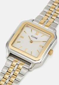 Gold- und silberne rechteckige Uhr mit einem weißen strukturierten Ziffernblatt, goldenen Stundenmarkierungen und einem gemischten Metallarmband. Verfügt über eine auffällige Krone.