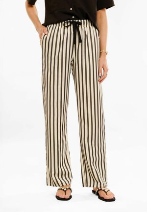 Femme portant un pantalon large à rayures verticales noires et beiges avec un cordon de serrage noir, un top noir et des sandales noires avec des accents en perles.