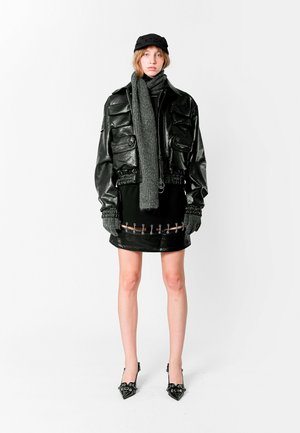 Chaqueta de cuero acolchada negra con bolsillos con cremallera, bufanda gris, vestido negro ajustado con acentos metálicos y tacones negros de punta.