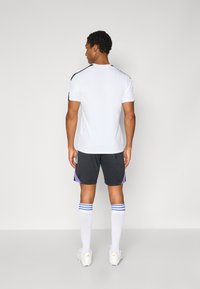T-shirt de sport blanche avec des accents noirs sur les épaules, associé à un short noir avec des détails violets et verts. Porté avec des chaussettes blanches rayées.