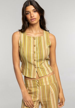 Mujer con top sin mangas color mostaza y pantalones a juego con rayas verticales blancas y marrones, de pie frente a un fondo liso.