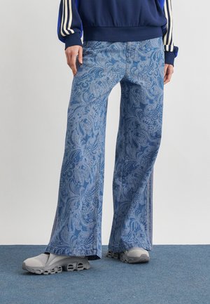 Person trägt blaue, weit geschnittene Hose mit Paisley-Muster, graue klobige Sneakers und einen dunkelblauen Pullover mit weißen Streifen an den Ärmeln.