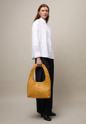 Donna con camicia bianca abbottonata e pantaloni neri che tiene una grande borsa in pelle color senape con borchie, su sfondo neutro.