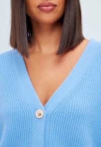 Jasnoniebieski sweter na drutach z dekoltem w serek, z pojedynczym okrągłym guzikiem z przodu i prążkowaną fakturą na całej tkaninie.