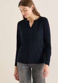 Granatowy top z długim rękawem, dekoltem w serek i plisowanym detalem z przodu. Wykonany z miękkiego materiału o gładkiej fakturze, zestawiony z szarymi jeansami.