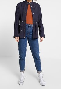 Giacca navy con colletto alto, bottoni a pressione metallici e vita regolabile indossata sopra un top color ruggine, abbinata a jeans blu e sneakers bianche.