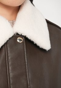 Jachetă din piele maro cu guler din blană albă, având un detaliu cu nasture auriu și o textură netedă. Focalizare pe zona decolteului și a gulerului.