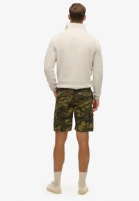 Sudadera de color claro con cuello alto, combinada con pantalones cortos cargo de camuflaje que cuentan con bolsillos laterales y un ajuste relajado, usados con zapatillas deportivas claras.