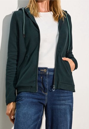 Frau trägt einen dunkelgrünen Kapuzenpullover mit Reißverschluss, weißes T-Shirt und blaue Jeans, steht mit einer Hand in der Hosentasche vor weißem Hintergrund.